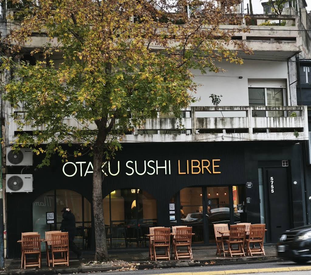 Sushi restaurant facade reading 'OTAKU SUSHI LIBRE' with wooden outdoor tables on the sidewalk and a leafy tree in front.